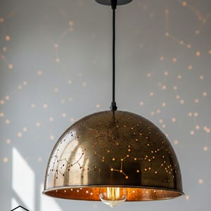 Puede incluir: Una lámpara colgante de color bronce con un patrón de constelación. La pantalla en forma de cúpula tiene estrellas y líneas recortadas, proyectando sombras en forma de estrella. Un cable negro y un soporte de techo sostienen la lámpara.