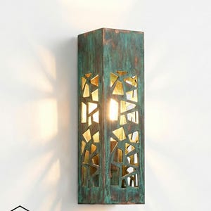 May include: A rectangular wall sconce with a weathered teal and bronze finish. The sconce features geometric cutouts that reveal a warm, golden light. The design includes the text "GHAZAL PEARLE" and a house logo.
