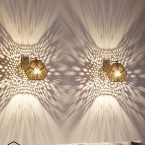 May include: Two gold-coloured, hexagonal wall sconces with intricate cut-out patterns, casting a decorative light and shadow effect on the wall. The sconces are mounted above a sofa with blue and brown cushions.