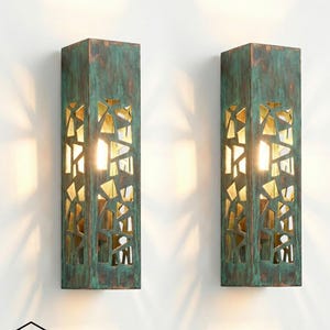 Könnte beinhalten: Zwei rechteckige Wandleuchten mit einer verwitterten teal- und bronzefarbenen Oberfläche. Jede Leuchte hat ein geometrisches Ausschnittdesign, das einen warmen, goldenen Innenraum freilegt. Die Leuchten sind an einer weißen Wand montiert und werfen weiches Licht.