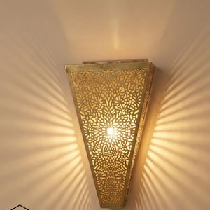May include: A gold-toned, triangular wall sconce featuring detailed floral cut-out patterns. The sconce is lit, creating a play of light and shadow on the wall. The brand logo "Ghazal Pearle" is displayed at the bottom.