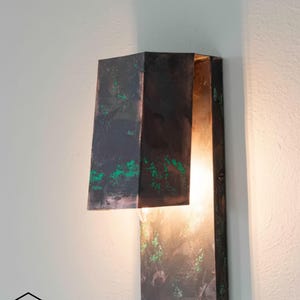 Peut inclure: Applique murale moderne avec une finition sombre et texturée et des accents verts. L'applique a une base rectangulaire et un abat-jour géométrique qui dirige la lumière vers le bas. Fixée sur un mur blanc.