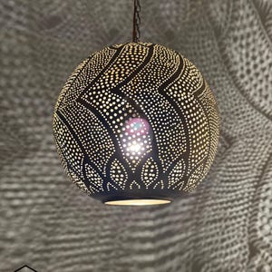 May include: A black, spherical pendant light with a patterned design of small, evenly spaced holes. The light hangs from a chain and casts intricate shadows. The light fixture has a warm glow, illuminating the detailed cut-out pattern.