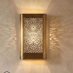 Könnte beinhalten: Rechteckige Wandleuchte aus Messing mit dekorativem, durchbrochenem Design. Das filigrane Muster lässt das Licht durchscheinen und erzeugt einen Muster-Effekt an der Wand. Die Leuchte ist an einer strukturierten, hellfarbigen Wand befestigt.