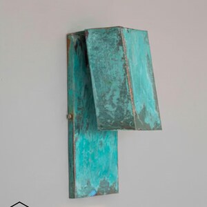 Peut inclure: Applique murale en métal patiné turquoise avec une plaque arrière rectangulaire et un abat-jour géométrique. Le luminaire présente des nuances de marron et de cuivre. Fixé sur un mur gris clair.