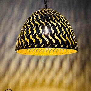 Puede incluir: Una lámpara colgante decorativa con exterior negro e interior amarillo. La luz tiene un patrón único en forma de onda cortado en la pantalla de metal. Cuelga de una cadena y un cable. La marca "GHAZAL PEARLE" es visible en la parte inferior.