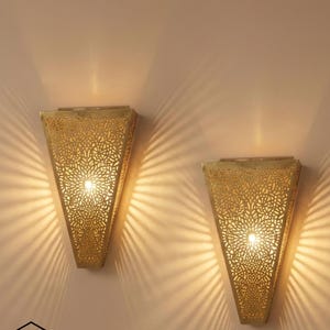 May include: Two gold-coloured, triangular wall sconces with intricate floral cut-out patterns. Each sconce emits a warm light, casting a patterned shadow on the wall. The sconces are mounted on a neutral-coloured wall.