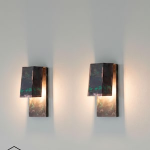 Peut inclure: Deux appliques murales modernes avec une finition sombre et texturée et des accents verts. Les appliques ont un design géométrique avec une base rectangulaire et un dessus évasé. La lumière brille du bas des appliques, créant une lueur chaleureuse. Elles sont montées sur un mur blanc.