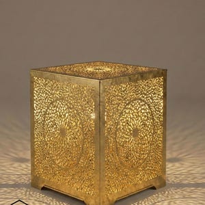 May include: A gold-colored, cube-shaped lantern with intricate, circular cut-out patterns. The lantern is illuminated from within, casting decorative shadows. The design features a floral motif, and the lantern sits on a flat surface.