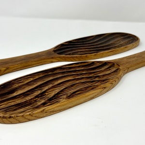 May include: Two wooden serving spoons with a textured, wave-like pattern carved into the bowl. The spoons are a warm brown color and have a handle with a hole for hanging. The spoons are likely for kitchen use.