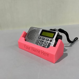 Peut inclure: Une radio numérique portable argentée avec une dragonne noire est placée dans un support en plastique rose personnalisé. L'avant du support présente le texte gravé "Your Name Here" dans une police sans empattement, offrant une base stable.