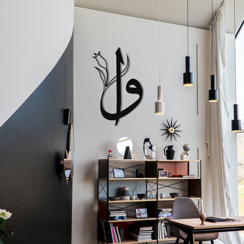 May include: A black metal wall art piece featuring Arabic calligraphy with a floral accent. The artwork is mounted on a white wall in a modern living space, alongside other decorative items and hanging lights.