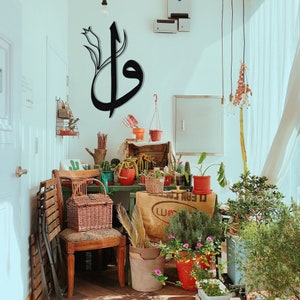 May include: A room filled with plants and decor. A large black metal wall art piece in Arabic calligraphy hangs on the wall. A wooden chair with a wicker basket sits in front of a table with various potted plants.