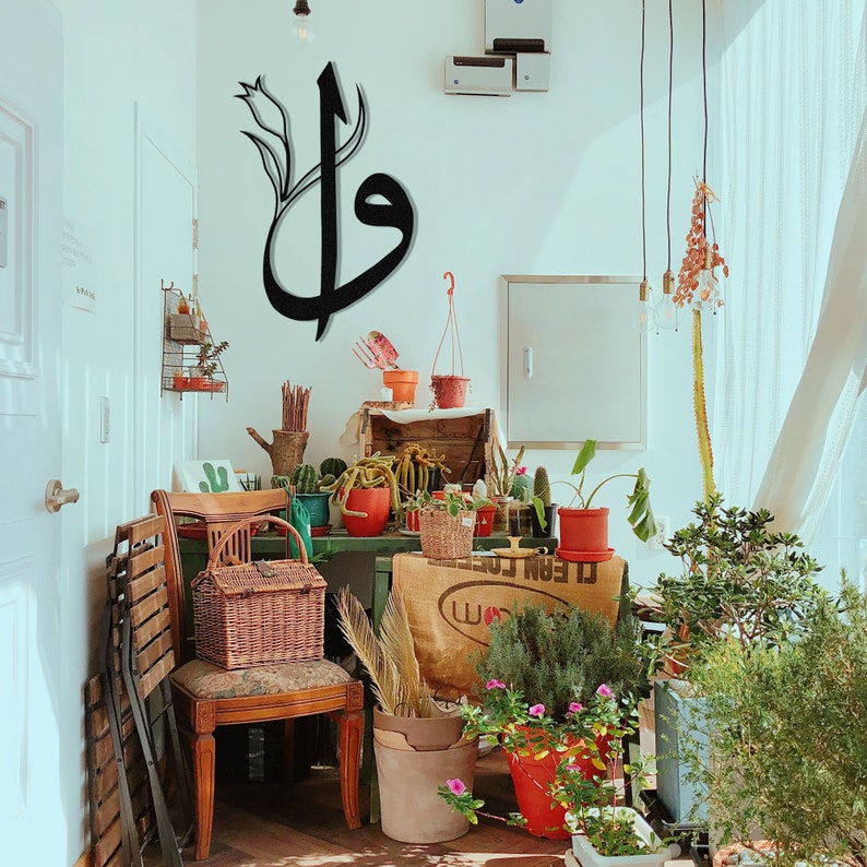 May include: A room filled with plants and decor. A large black metal wall art piece in Arabic calligraphy hangs on the wall. A wooden chair with a wicker basket sits in front of a table with various potted plants.