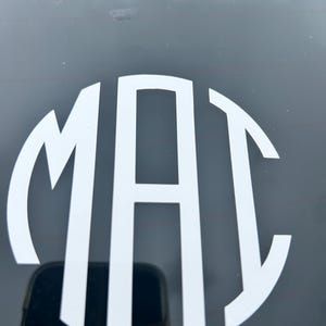 May include: A white vinyl decal featuring the letters M, A, and C in a circular monogram style. The letters are arranged with the central A being the tallest, flanked by the M and C, creating a cohesive, rounded design applied to a dark surface.