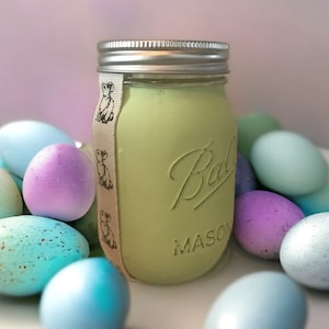 Pode incluir: Um frasco de vidro verde com tampa prateada e uma fita branca com silhuetas de coelhos pretos. O frasco está rodeado de ovos de Páscoa em tons pastel.