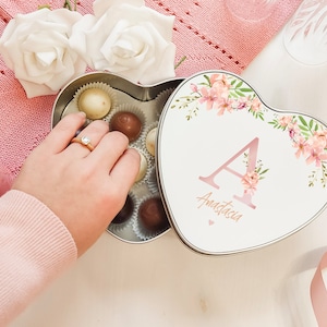 Può includere: Una scatola di latta a forma di cuore con un design floreale e il nome "Anastasia" e la lettera "A" in rosa. La scatola è aperta e piena di cioccolatini.