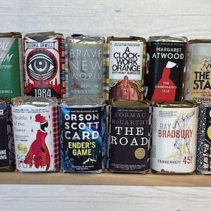 Può includere: Una collezione di 12 saponi in miniatura a forma di libro, ognuno con un diverso design di copertina. I saponi sono disposti su due file su uno scaffale di legno. Le copertine dei libri includono titoli come "La fattoria degli animali", "1984", "Il mondo nuovo", "Arancia meccanica", "Il racconto dell'ancella", "La torre nera", "Hunger Games", "Parable of the Sower", "Il gioco di Ender", "La strada", "Fahrenheit 451", e "Il donatore".