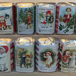 May include: Twelve small, rectangular, silver-wrapped soaps with Christmas-themed illustrations. Each soap features a different image of children in vintage clothing, including a girl with a red dress and a boy with a blue coat. The soaps have a plaid pattern on the sides and are arranged in two rows of six.