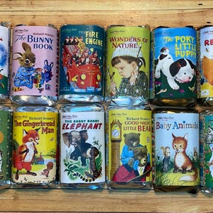 May include: A collection of vintage-style tin cans featuring colorful illustrations from children's books. Titles include "Tawny Lion," "The Bunny Book," "Fire Engine," and "The Poky Little Puppy." The cans are arranged on a wooden surface.