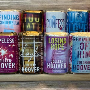 May include: A collection of 12 paperback books by Colleen Hoover. The books are arranged in a row on a wooden shelf. The book titles include "It Starts With Us", "Finding Cinderella", "Ugly Love", "It Ends With Us", "Hopeless", "Layla", "Losing Hope", "Reminder of Him", "Regretting You", "Confess", "Too Late", and "All Your Perfects".