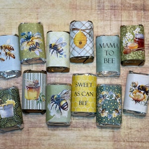 May include: Assortment of small, wrapped chocolate bars with bee-themed designs. The wrappers feature bees, honeycombs, jars of honey, and floral patterns. Some wrappers have text, including "Mama to Bee", "Sweet as Can Bee", and "Bee Happy".