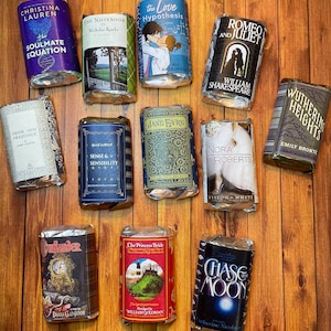 May include: A collection of 12 wrapped book-shaped soaps, each featuring a different book cover design. The soaps are arranged in three rows of four on a wooden surface. The book covers include titles such as "The Notebook", "Pride and Prejudice", "Sense and Sensibility", "Jane Eyre", "The Love Hypothesis", "Romeo and Juliet", "Wuthering Heights", "The Princess Bride", "Outlander", and "Chase the Moon".