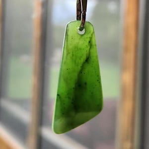 Könnte beinhalten: Ein grüner Jade-Anhänger mit silberfarbenem Verschluss. Der Anhänger hat eine hellgrüne Farbe mit dunkleren grünen und schwarzen Einschlüssen. Die Form ist ein abgerundetes Trapez. Der Anhänger hängt vor einem verschwommenen Hintergrund.