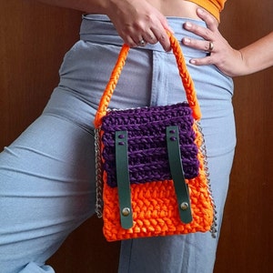 Op de afbeelding: Een klein, gehaakt handtasje met een paars en oranje ontwerp. De tas heeft een lange band en een zilveren kettingdetail.