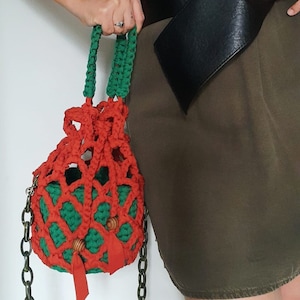Pode incluir: Uma bolsa de crochê vermelha e verde com contas de madeira e uma longa alça de corrente. A bolsa está pendurada no braço de uma pessoa que está usando uma saia marrom.