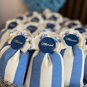 以下が含まれることがあります： 青と白のストライプの布製の袋に、白いレースと「Marsel」と書かれた青い丸いタグが付いています。袋は銀色のワイヤーバスケットに並べられています。