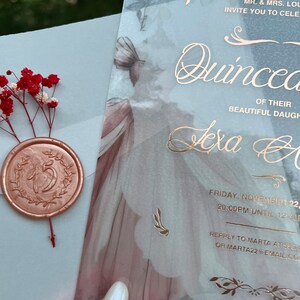 May include: A translucent blue and white invitation with gold lettering for a Quincea&ntilde;era. The invitation features a floral design and the text "MR. & MRS. LOU INVITE YOU TO CELEBRATE THE QUINCEA&Ntilde;ERA OF THEIR BEAUTIFUL DAUGHTER TEXA A FRIDAY, NOVEMBER 22, 20:00PM UNTIL 12:30 REPPLY TO MARTA AT 555 OR MARTA22@EMAIL.COM".