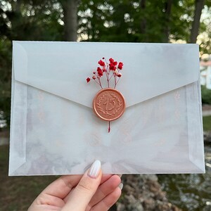 May include: A white envelope with a wax seal featuring the number "15" in a wreath design. The seal is decorated with a small bouquet of red flowers.