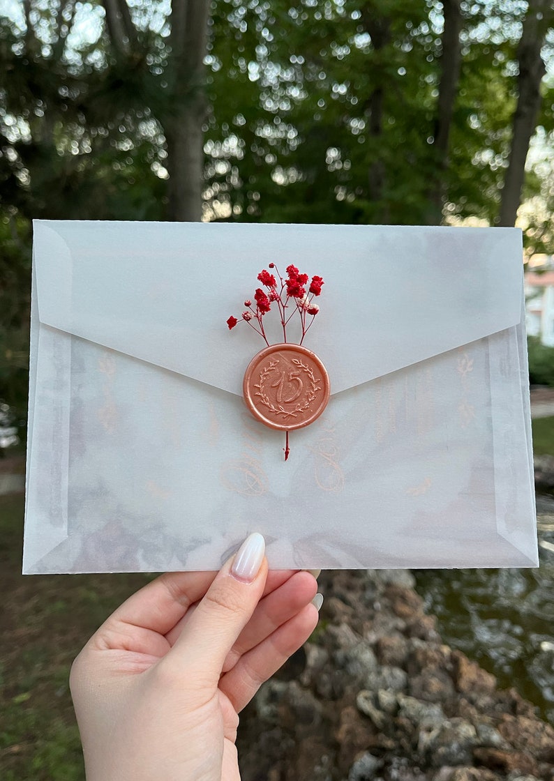 May include: A white envelope with a wax seal featuring the number "15" in a wreath design. The seal is decorated with a small bouquet of red flowers.
