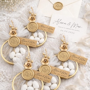 Könnte beinhalten: Hochzeitsgeschenke mit goldfarbenen, kreisförmigen Mesh-Körben, gefüllt mit weißen Süßigkeiten, verziert mit goldenen Blumenakzenten und Etiketten mit der Aufschrift "Alexa & Max 26.03.2026." Eine Einladung und ein Umschlag mit goldenem Siegel sind ebenfalls vorhanden.