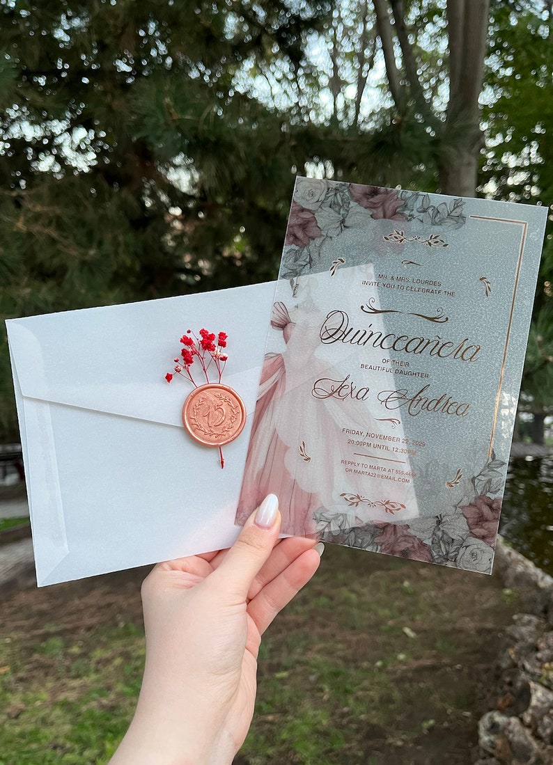 May include: A clear plastic invitation with a floral design and gold accents. The invitation is for a Quincea&ntilde;era, a celebration of a girl's 15th birthday. The text on the invitation reads "M&Mrs. Lourdes invite you to celebrate the Quincea&ntilde;era of their beautiful daughter Alexa Andrea. Friday, November 28, 2029, 20:00pm until 12:20am. Reply to Marta at 555-555-5555 or MartazzEmail.com."