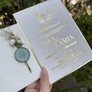 Op de afbeelding: Elegante trouwuitnodiging met goudfolie letters en accenten. De uitnodiging toont de namen "SAFA LAMIA" en evenementdetails. Een envelop met een turquoise waszegel en bloemversieringen is inbegrepen.