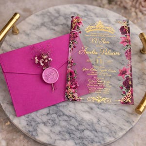 May include: A magenta invitation suite featuring a clear acrylic invitation with gold script and floral accents. A matching magenta envelope is sealed with a pink wax seal and small floral arrangement. The suite rests on a marble tray.