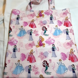 Może przedstawiać: R&oacute;żowa torba typu tote z powtarzającym się wzorem księżniczek Disneya, w tym Kopciuszka, Ariel, Belle i Mulan. Księżniczki są ubrane w swoje charakterystyczne suknie i otoczone r&oacute;żowymi kwiatami i chmurami.