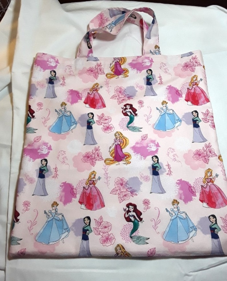 Może przedstawiać: R&oacute;żowa torba typu tote z powtarzającym się wzorem księżniczek Disneya, w tym Kopciuszka, Ariel, Belle i Mulan. Księżniczki są ubrane w swoje charakterystyczne suknie i otoczone r&oacute;żowymi kwiatami i chmurami.