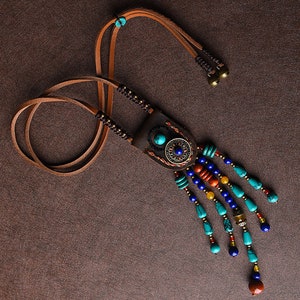 Puede incluir: Un collar largo de cuero con un colgante turquesa y de latón y una franja de cuentas de colores. Las cuentas son turquesa, azul, naranja, rojo y amarillo.