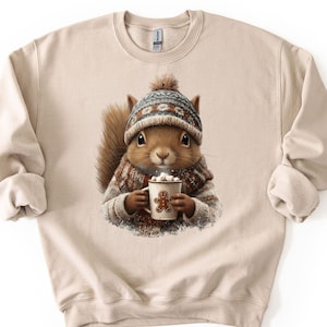 May include: Beige sweatshirt featuring a cute squirrel wearing a winter hat and scarf, holding a mug of hot chocolate with marshmallows. The squirrel has a gingerbread man design on the mug. The sweatshirt has long sleeves and a crew neck.