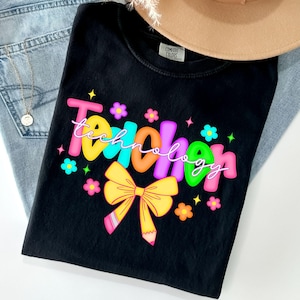 May include: Black t-shirt with the word "Teacher" in colorful bubble letters and the word "Technology" in a cursive font. The design includes a yellow bow, a pencil, and floral accents. The shirt is paired with blue jeans and a tan hat.