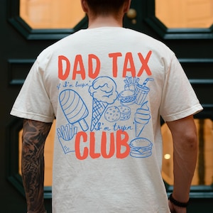 May include: An ivory short-sleeve t-shirt featuring a graphic on the back with the red text DAD TAX CLUB and blue line art illustrations of snacks including ice cream, fries, and burgers with the text if I'm buyin' I'm tryin'.