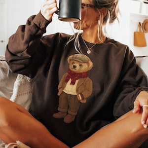 May include: A dark brown sweatshirt with an embroidered teddy bear design. The bear wears a cap, scarf, and jacket, and holds a book. The person is holding a black mug. The sweatshirt is a casual, comfortable style.