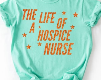 May include: A light turquoise t-shirt with rolled-up sleeves. The shirt features the text "THE LIFE OF A HOSPICE NURSE" in orange glitter lettering, along with orange star graphics. The shirt is made of a soft material.