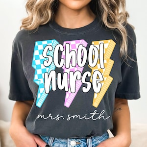 May include: Dark gray t-shirt with the words "SCHOOL NURSE" in white, with three lightning bolt designs in blue and white checkered, pink, and yellow. The name "mrs. smith" is written below the text.