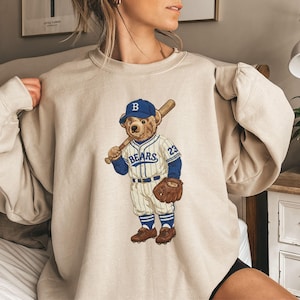 May include: Beige sweatshirt with a baseball-playing bear illustration. The bear wears a blue cap with a "B" and a baseball uniform with "BEARS" and the number "23". The bear holds a bat and a glove. The sweatshirt has a crew neck.