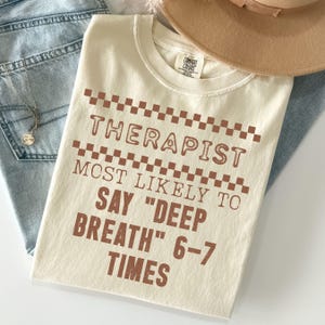 May include: Cream-colored t-shirt with the text "THERAPIST MOST LIKELY TO SAY 'DEEP BREATH' 6-7 TIMES" in brown letters. The shirt is folded, with a brown hat and blue jeans in the background.