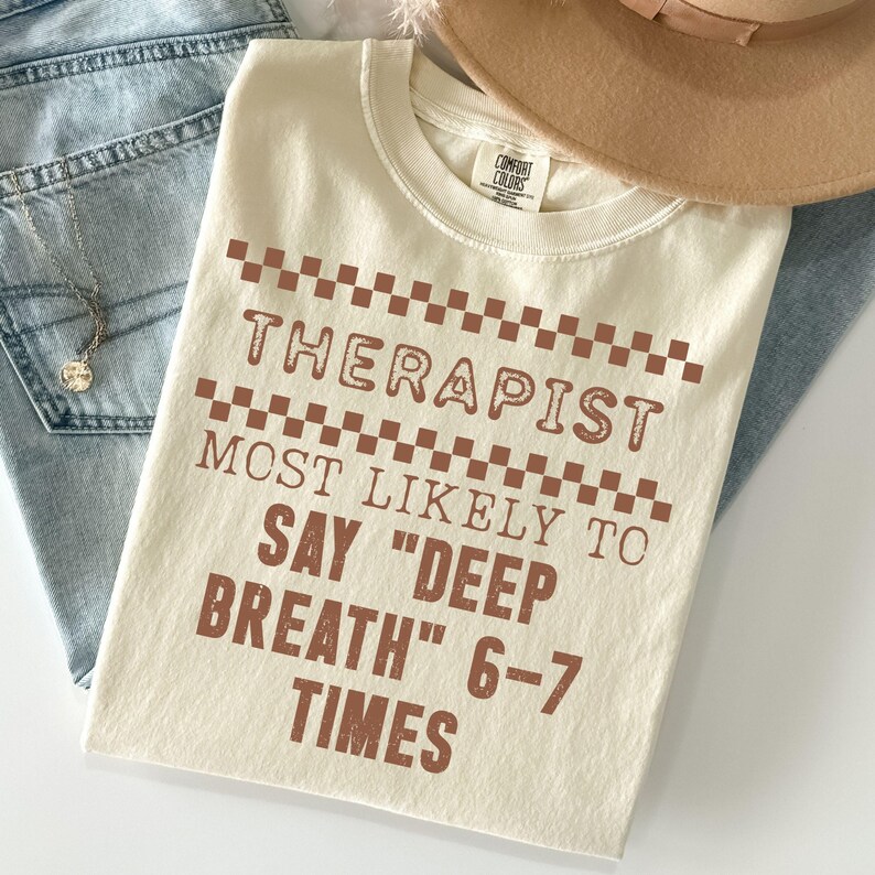 May include: Cream-colored t-shirt with the text "THERAPIST MOST LIKELY TO SAY 'DEEP BREATH' 6-7 TIMES" in brown letters. The shirt is folded, with a brown hat and blue jeans in the background.